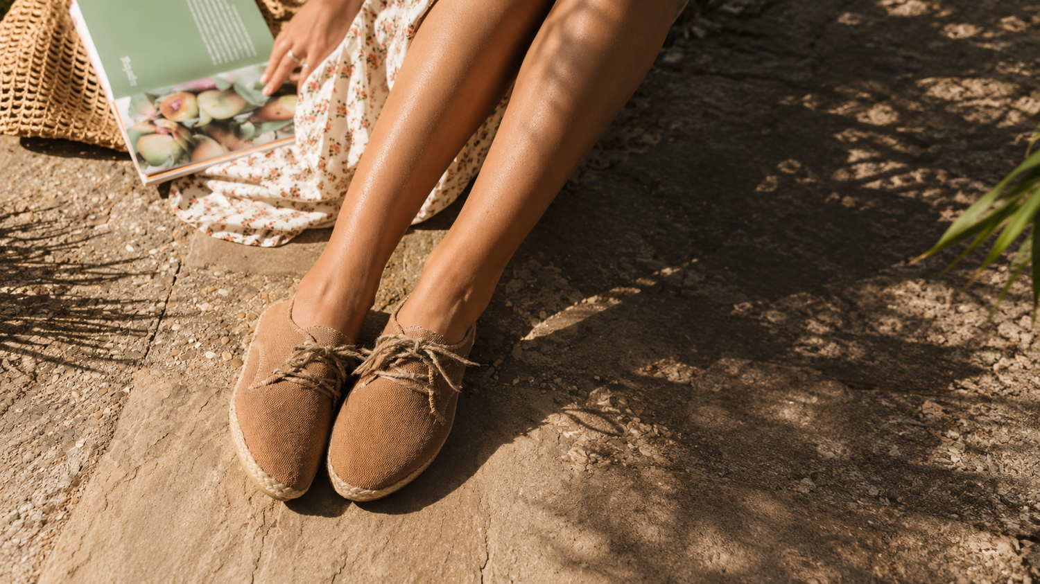 A person sitting outdoors on a stone surface wearing tan suede lace-up shoes, with a patterned skirt, a woven bag, and a magazine nearby in warm natural light.