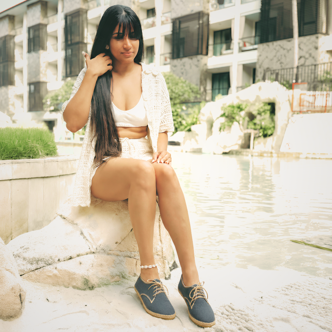 A woman with long black hair sits on a stone ledge next to a swimming pool, wearing a white crop top, white shorts, a white open-front cardigan, and navy blue lace-up shoes.