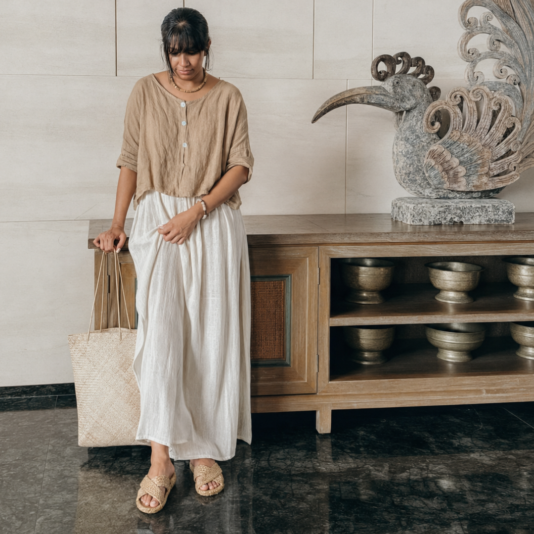 A woman stands indoors wearing a loose beige short-sleeve top and a long white skirt, holding a woven tote bag beside a wooden cabinet with decorative bowls and a large bird sculpture.