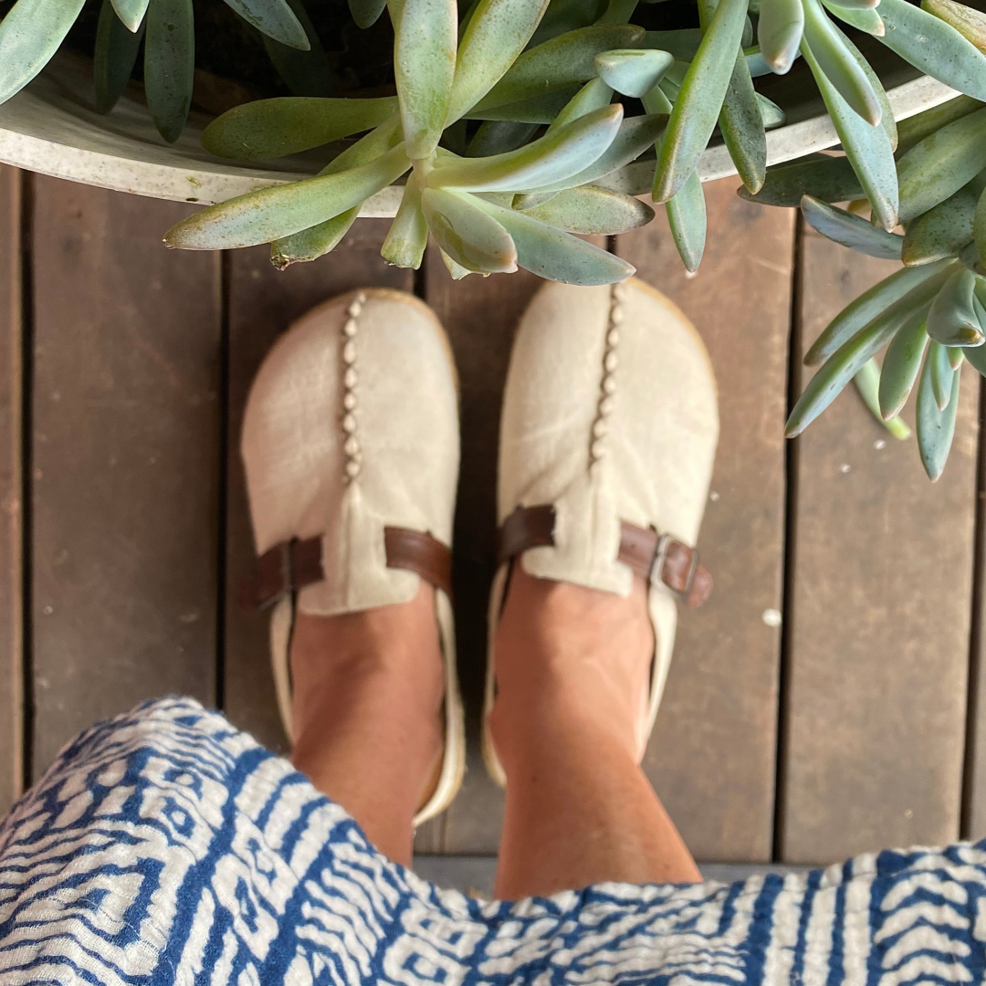 A top-down view of a person wearing a pair of off-white canvas mule shoes with brown straps, standing on a wooden deck with a green succulent plant in the foreground.