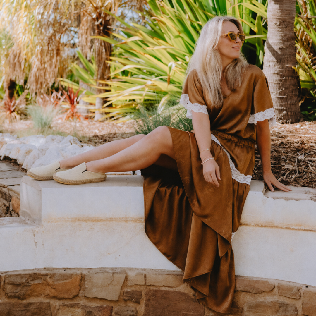 A woman wearing a brown satin-like dress with white lace trim sits on a white stone ledge in an outdoor garden setting with palm trees.