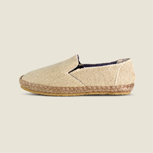 A single slip-on espadrille-style shoe in a textured beige fabric upper with a braided jute trim and a tan outsole, shown in side profile against a light beige background.
